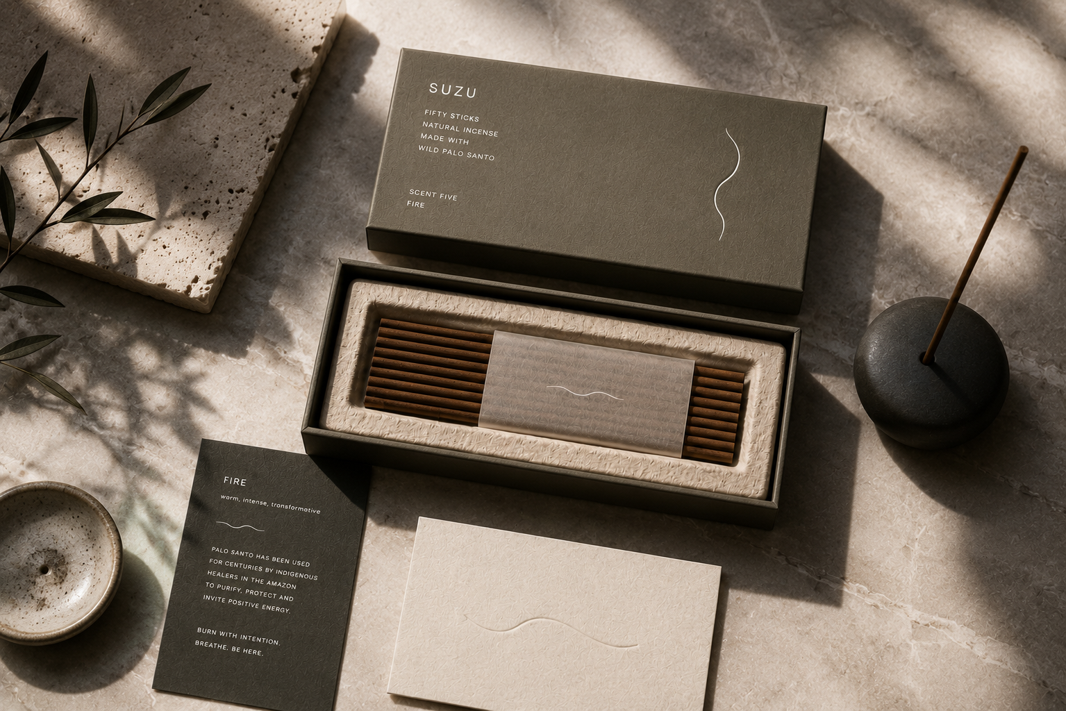 Suzu Fire incense box with palo santo sticks, ceramic holder, and olive branch on warm stone